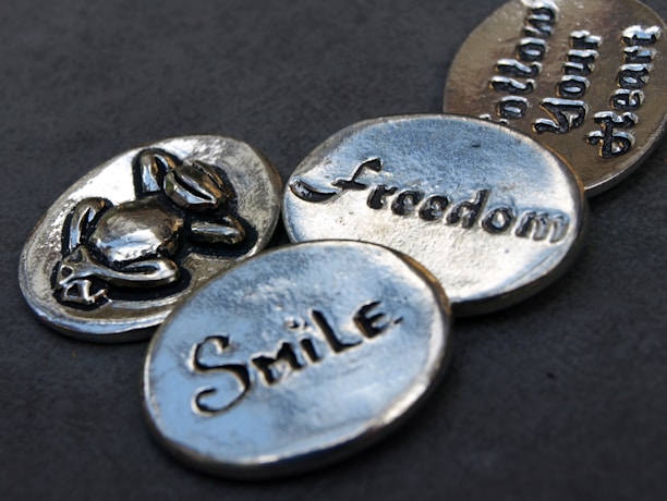 Four silver metallic tokens lie on a dark gray surface. Each token has a word or an image engraved on it. One token has a symbol of a turtle, another says 'Freedom', the third reads 'Smile', and the fourth one says 'Follow Your Heart'. The tokens have a shiny, polished appearance with a slightly worn texture.