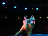 Two dancers perform on a dimly lit stage, illuminated by blue spotlights. They are positioned closely, with one dancer holding the other. The background is dark, with a few glowing windows visible, suggesting an indoor venue or theater.