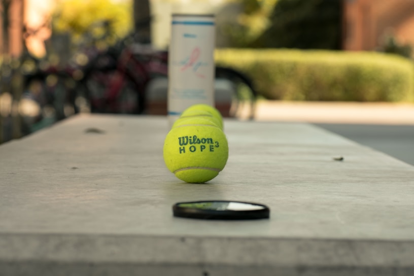 A tennis ball branded with 'Wilson Hope' is centered in focus on a blurred background that appears to be outdoors. There is a faintly visible object in the foreground, possibly a lens cap or a similar small item. The background is out of focus, with traces of greenery and what looks like a can or container.