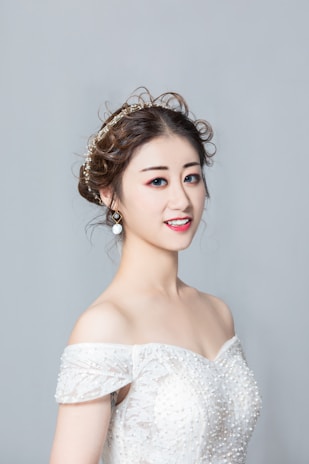 A woman is wearing an off-the-shoulder white dress with intricate beadwork and patterns. Her hair is styled in an elegant updo with curls, adorned with a decorative hairpiece. She is wearing makeup, including red lipstick and eye makeup, and is smiling slightly.