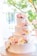 A three-tiered wedding cake adorned with delicate pink and white roses is set on an intricately decorated stand. The background features blurred greenery and soft, warm lighting, creating a serene and elegant atmosphere.