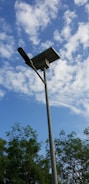 A tall streetlight with a solar panel on top is positioned against a background of a partly cloudy blue sky. Trees with green foliage are visible at the bottom of the image.