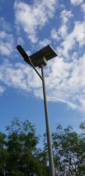 A tall streetlight with a solar panel on top is positioned against a background of a partly cloudy blue sky. Trees with green foliage are visible at the bottom of the image.
