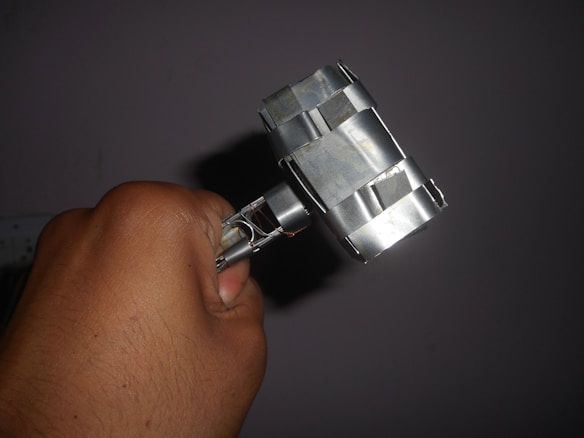 A hand holding a homemade hammer-like object made of metallic components, featuring a blocky head and handle secured with metal strips.