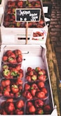 Several plastic containers filled with ripe red strawberries are stacked inside a cardboard box. A small chalkboard sign displays the price of the strawberries. The setup is on a wooden market stall, suggesting a fresh market or grocery setting.