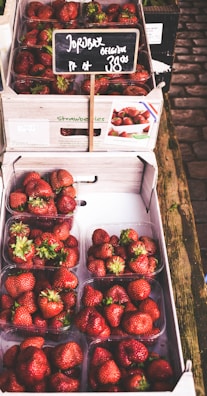 Several plastic containers filled with ripe red strawberries are stacked inside a cardboard box. A small chalkboard sign displays the price of the strawberries. The setup is on a wooden market stall, suggesting a fresh market or grocery setting.