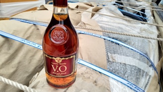 A bottle of brandy with a maroon and gold label is placed on a tan paper surface. The bottle is surrounded by packaging material including bubble wrap and ropes.