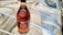 A bottle of brandy with a maroon and gold label is placed on a tan paper surface. The bottle is surrounded by packaging material including bubble wrap and ropes.