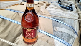 A bottle of brandy with a maroon and gold label is placed on a tan paper surface. The bottle is surrounded by packaging material including bubble wrap and ropes.