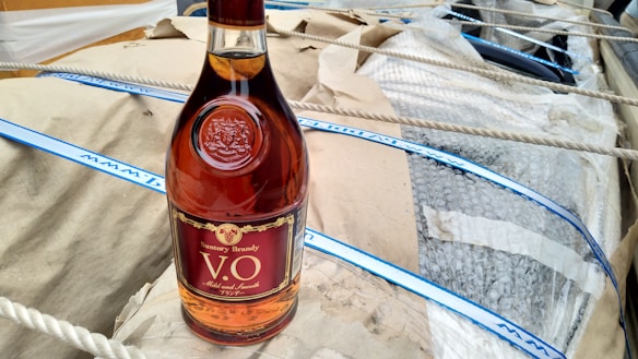 A bottle of brandy with a maroon and gold label is placed on a tan paper surface. The bottle is surrounded by packaging material including bubble wrap and ropes.