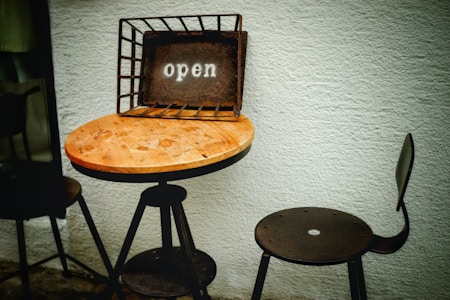 A round wooden table with a metal stand is placed against a white textured wall. On top of the table is an open sign within a rustic metal frame, and two chairs are positioned nearby, one directly facing and the other partially visible.