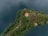 An aerial view of a small, lush green island surrounded by dark blue water, featuring a few structures with a prominent building having an orange roof. The island is dense with trees and vegetation, with footpaths connecting the buildings, conveying a serene and secluded atmosphere.