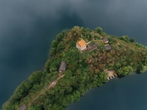 An aerial view of a small, lush green island surrounded by dark blue water, featuring a few structures with a prominent building having an orange roof. The island is dense with trees and vegetation, with footpaths connecting the buildings, conveying a serene and secluded atmosphere.