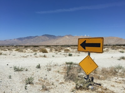 A desert landscape with sparse vegetation and a backdrop of mountains under a clear blue sky. In the foreground, two road signs are visible; one is a yellow diamond, and the other is a left-pointing yellow arrow.
