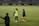 Two people are on a soccer field, wearing sports attire. One person is wearing a green vest, and the other is in a blue jacket and white shorts. The setting appears to be during a sports event, with adverts in the background and a partially filled stadium.