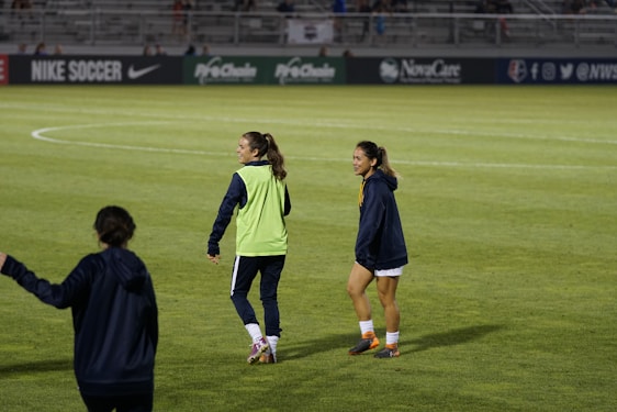 Two people are on a soccer field, wearing sports attire. One person is wearing a green vest, and the other is in a blue jacket and white shorts. The setting appears to be during a sports event, with adverts in the background and a partially filled stadium.