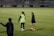 Two people are on a soccer field, wearing sports attire. One person is wearing a green vest, and the other is in a blue jacket and white shorts. The setting appears to be during a sports event, with adverts in the background and a partially filled stadium.