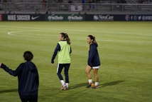 Two people are on a soccer field, wearing sports attire. One person is wearing a green vest, and the other is in a blue jacket and white shorts. The setting appears to be during a sports event, with adverts in the background and a partially filled stadium.