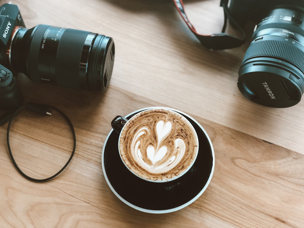 Cappuccino auf Keramikbecher zwischen zwei Sony und Tanron DSLR-Kameras auf brauner Holzoberfläche