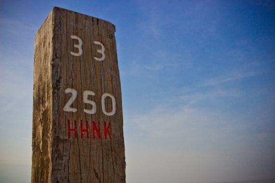 A wooden post stands vertically with numbers 33 and 250 engraved in white and the letters HHNK in red. The background features a clear blue sky.