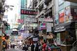 A bustling urban street scene filled with numerous colorful signage written in various languages, including Chinese and English. The street is lined with multi-story buildings featuring shops and restaurants, with people walking along the narrow road. Some of the shops include a tailor and a café, and vehicles are parked along the street.