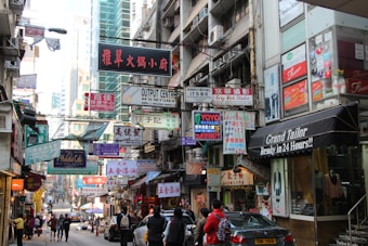 A bustling urban street scene filled with numerous colorful signage written in various languages, including Chinese and English. The street is lined with multi-story buildings featuring shops and restaurants, with people walking along the narrow road. Some of the shops include a tailor and a caf&eacute;, and vehicles are parked along the street.