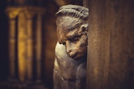 A close-up view of a stone sculpture featuring a contemplative face with a furrowed brow, casting a shadow against an ancient architectural background.