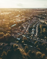 An aerial view of a sprawling town featuring a mix of densely-packed residential buildings and lush green spaces. The scene is bathed in warm, golden sunlight, casting long shadows from trees lining a winding river in the foreground. Roads weave through neighborhoods, connecting rows of houses with red-tiled roofs.