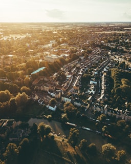 An aerial view of a sprawling town featuring a mix of densely-packed residential buildings and lush green spaces. The scene is bathed in warm, golden sunlight, casting long shadows from trees lining a winding river in the foreground. Roads weave through neighborhoods, connecting rows of houses with red-tiled roofs.