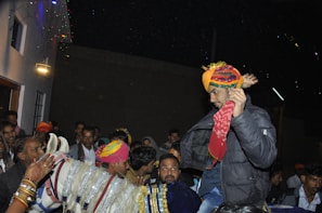 A man wearing a colorful turban and dark jacket is sitting on a horse, surrounded by a crowd of people. The scene appears to be part of a festive or cultural event taking place at night, with decorations and lights visible.