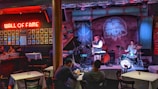 A cozy jazz bar features live music with a small band performing on stage. The room is warmly lit with red and blue lights, highlighting the musicians: a saxophonist, a drummer, and a bassist. Patrons are seated at tables enjoying the performance. A 'Wall of Fame' with framed photos is prominently displayed on the left wall.