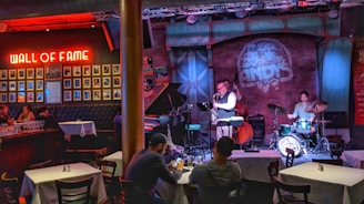 A cozy jazz bar features live music with a small band performing on stage. The room is warmly lit with red and blue lights, highlighting the musicians: a saxophonist, a drummer, and a bassist. Patrons are seated at tables enjoying the performance. A 'Wall of Fame' with framed photos is prominently displayed on the left wall.