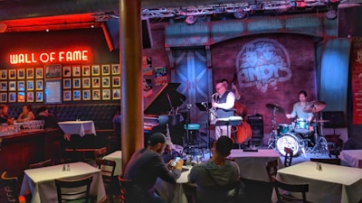 A cozy jazz bar features live music with a small band performing on stage. The room is warmly lit with red and blue lights, highlighting the musicians: a saxophonist, a drummer, and a bassist. Patrons are seated at tables enjoying the performance. A 'Wall of Fame' with framed photos is prominently displayed on the left wall.