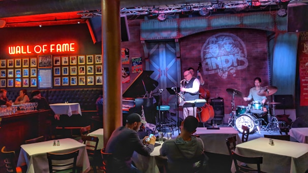 A cozy jazz bar features live music with a small band performing on stage. The room is warmly lit with red and blue lights, highlighting the musicians: a saxophonist, a drummer, and a bassist. Patrons are seated at tables enjoying the performance. A 'Wall of Fame' with framed photos is prominently displayed on the left wall.