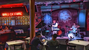 A cozy jazz bar features live music with a small band performing on stage. The room is warmly lit with red and blue lights, highlighting the musicians: a saxophonist, a drummer, and a bassist. Patrons are seated at tables enjoying the performance. A 'Wall of Fame' with framed photos is prominently displayed on the left wall.