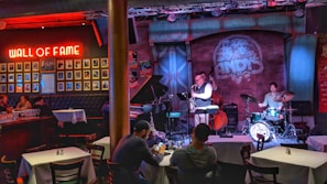 A cozy jazz bar features live music with a small band performing on stage. The room is warmly lit with red and blue lights, highlighting the musicians: a saxophonist, a drummer, and a bassist. Patrons are seated at tables enjoying the performance. A 'Wall of Fame' with framed photos is prominently displayed on the left wall.