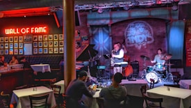 A cozy jazz bar features live music with a small band performing on stage. The room is warmly lit with red and blue lights, highlighting the musicians: a saxophonist, a drummer, and a bassist. Patrons are seated at tables enjoying the performance. A 'Wall of Fame' with framed photos is prominently displayed on the left wall.