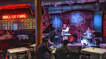 A cozy jazz bar features live music with a small band performing on stage. The room is warmly lit with red and blue lights, highlighting the musicians: a saxophonist, a drummer, and a bassist. Patrons are seated at tables enjoying the performance. A 'Wall of Fame' with framed photos is prominently displayed on the left wall.
