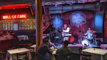 A cozy jazz bar features live music with a small band performing on stage. The room is warmly lit with red and blue lights, highlighting the musicians: a saxophonist, a drummer, and a bassist. Patrons are seated at tables enjoying the performance. A 'Wall of Fame' with framed photos is prominently displayed on the left wall.