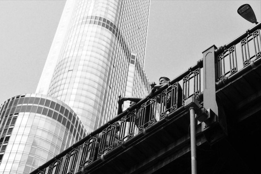 Fotografía en escala de grises de ángulo bajo de dos hombres que apoyan ambos brazos en un balaustre al aire libre junto a un edificio de gran altura durante el día