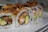 A close-up of sushi rolls filled with crispy tempura shrimp, avocado, and topped with fried onions and a sprinkle of tempura flakes. The rolls are neatly arranged on a white rectangular plate, highlighting the textures and colors of the ingredients.