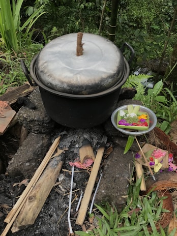A large metal pot with a wooden handle is resting on a traditional outdoor cook stove made of stones and wood. Surrounding the pot is a lush green environment with various plants and bamboo. A metal tray containing colorful flowers and offerings is placed nearby, adding a hint of cultural or ceremonial context.