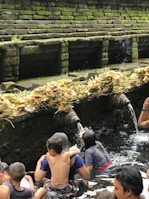 A group of people are engaged in a ritual bath at a sacred water temple. Stone spouts carved into the temple wall are pouring water, and numerous offerings made of palm leaves and flowers are placed on the ledge above the water source. The stone walls are covered with moss, adding a natural green hue to the scene.