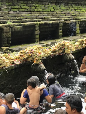 A group of people are engaged in a ritual bath at a sacred water temple. Stone spouts carved into the temple wall are pouring water, and numerous offerings made of palm leaves and flowers are placed on the ledge above the water source. The stone walls are covered with moss, adding a natural green hue to the scene.