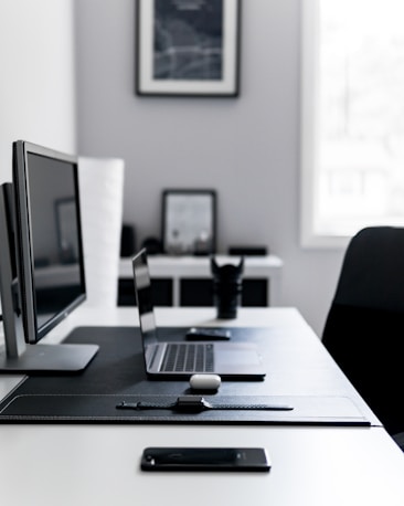A minimalist desk setup featuring a laptop, desktop monitor, a smartphone, and a wireless earbud case on a clean black desk pad. The background shows a framed abstract artwork and a blurred window.