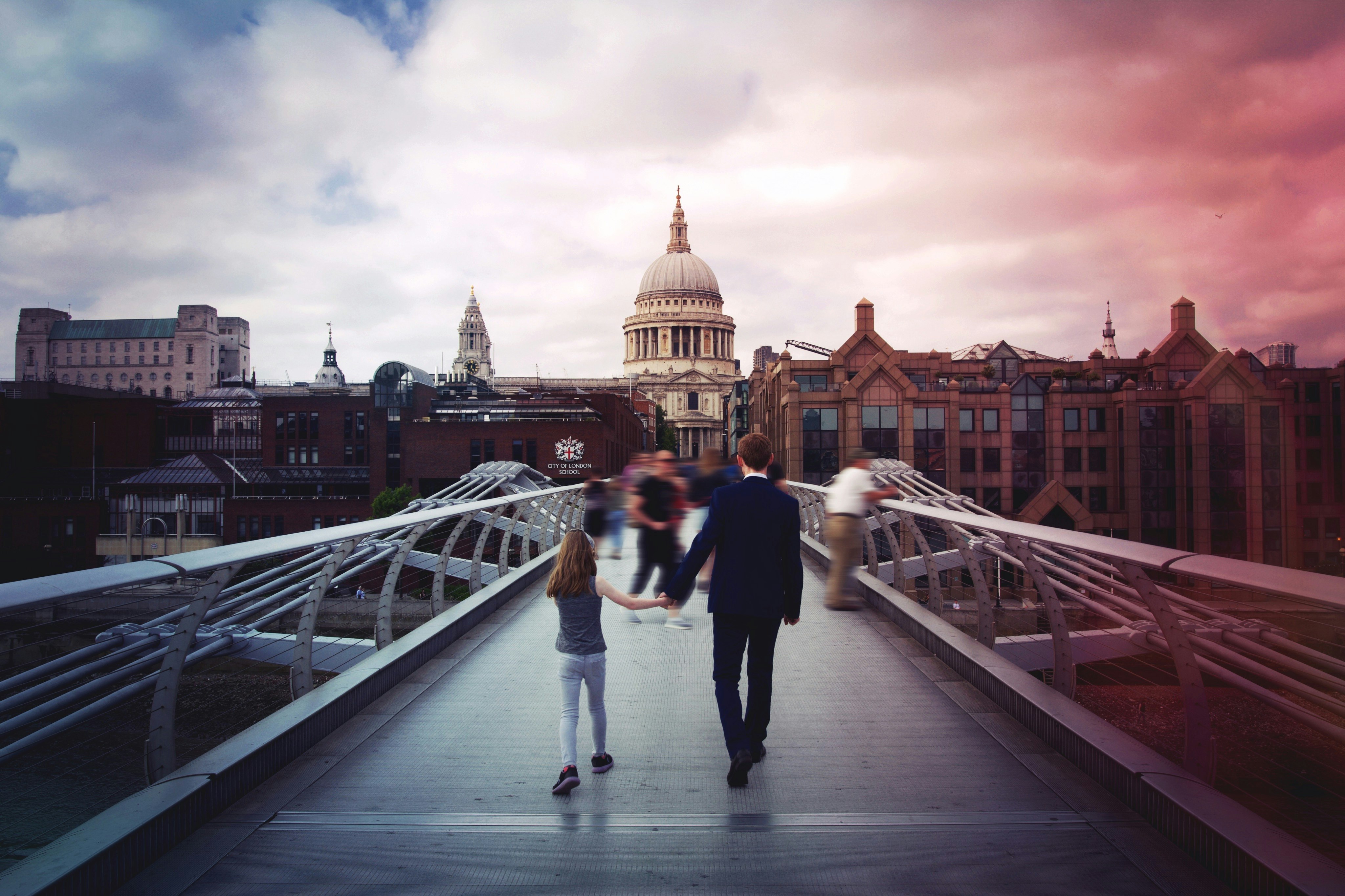 man and girl holding hands while walking on bridge in front of concrete dome building
