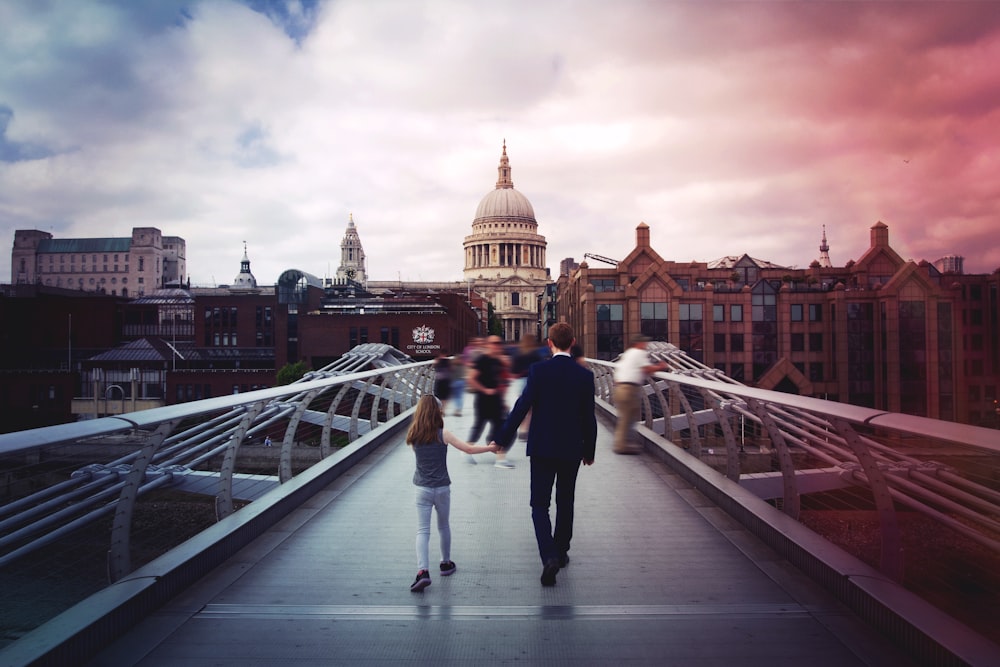 uomo e ragazza che si tengono per mano mentre camminano sul ponte di fronte all'edificio della cupola di cemento