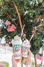 two persons holding Joe's frappes plastic cups near tree