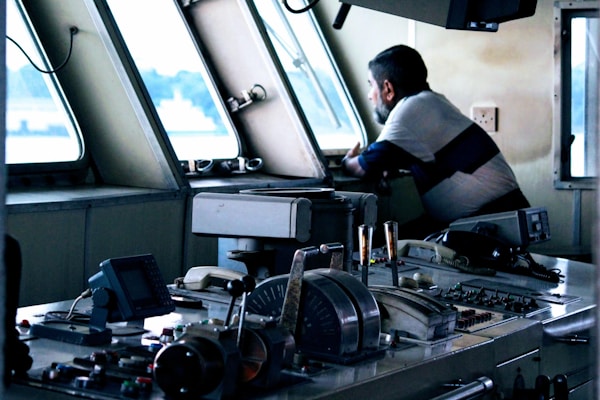 A man is leaning on a control panel inside a boat or ship's bridge, looking out of the window. The setting features various navigational instruments and controls, with a focus on levers, buttons, and communication devices. The overall ambiance suggests a maritime environment.