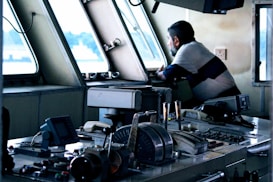 A man is leaning on a control panel inside a boat or ship's bridge, looking out of the window. The setting features various navigational instruments and controls, with a focus on levers, buttons, and communication devices. The overall ambiance suggests a maritime environment.